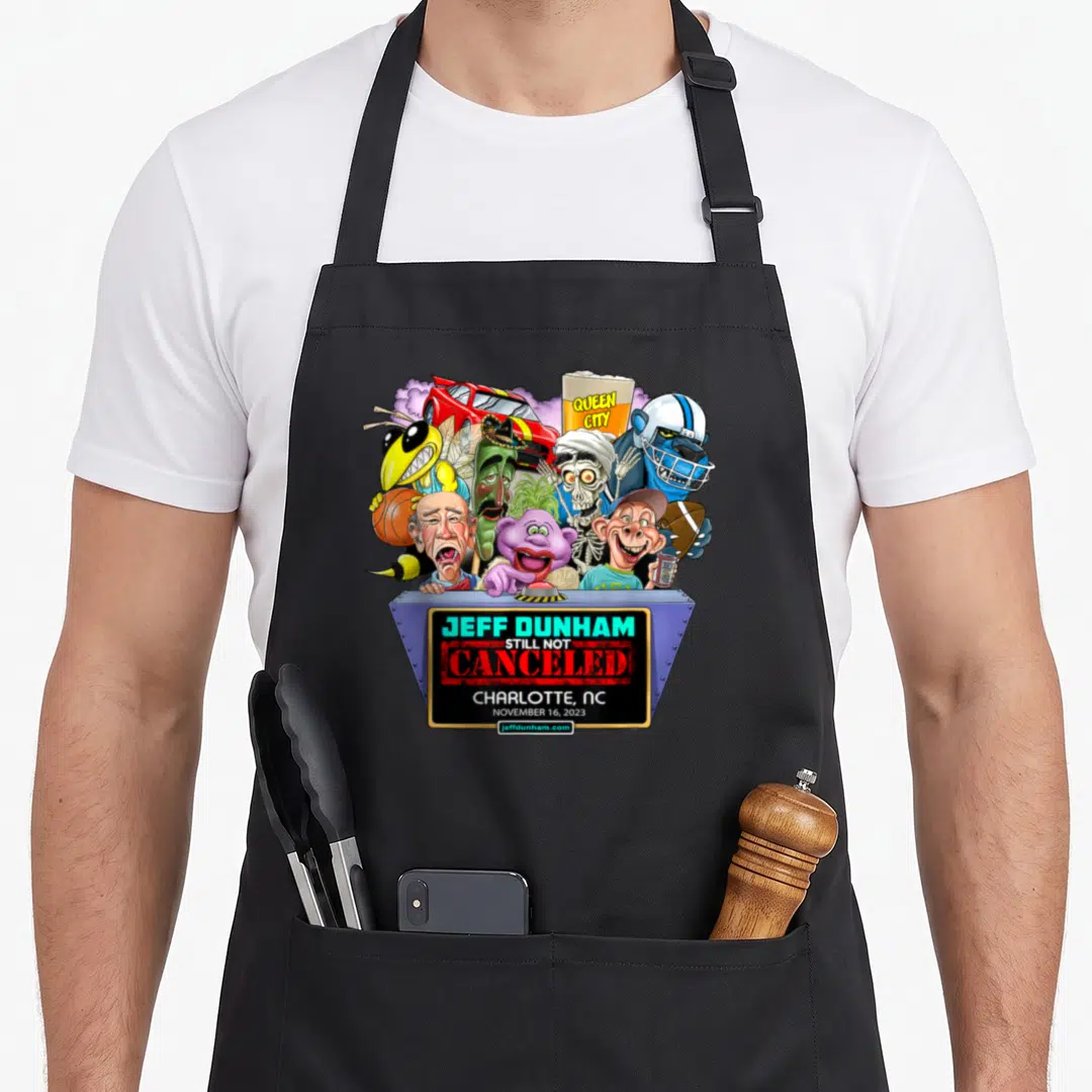 Dunham charlotte apron - Cartoon puppets above grey podium with Jeff Dunham Still Not Canceled text — shown on a black cotton canvas apron against a white background