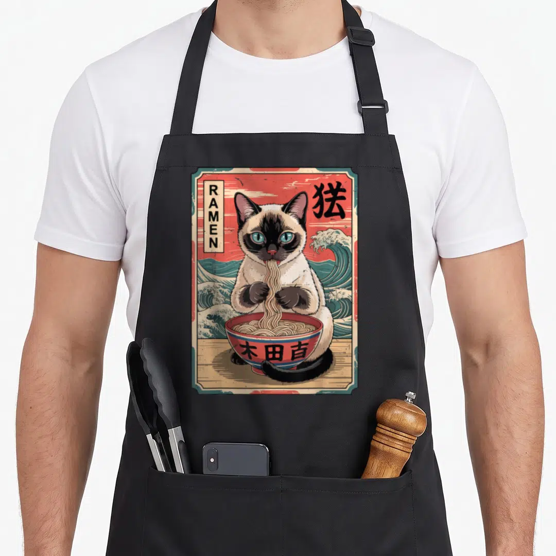 Eating cute cat aprons - Siamese cat eating ramen noodles from red bowl with retro waves and RAMEN text — shown on a black cotton canvas apron against a white background