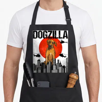 Dogzilla rhodesian ridgeback apron - Giant brown dog sitting before red sun and city skyline with helicopters — shown on a black cotton canvas apron against a white background