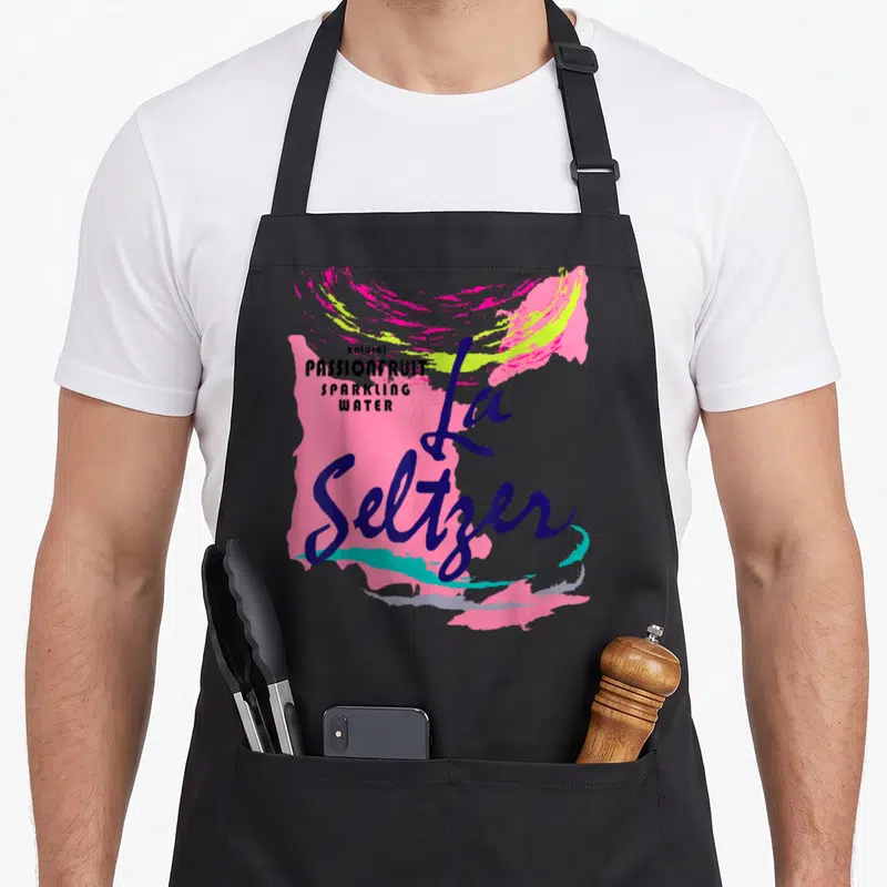 Passionfruit halloween apron - La Seltzer script text over pink and turquoise paint splashes with stacked flavor details — shown on a black cotton canvas apron against a white background