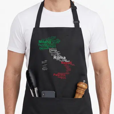 Travel italy apron - Italian city names forming a map shape in green white and red text — shown on a black cotton canvas apron against a white background