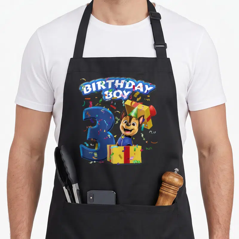 Cool chase birthday apron - BIRTHDAY BOY text, blue number 3, dog in yellow gift box with confetti — shown on a black cotton canvas apron against a white background