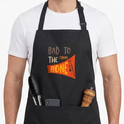 Pun trombone quotes apron - Bad to the trom bone text with orange banner and cartoon trombone illustration — shown on a black cotton canvas apron against a white background