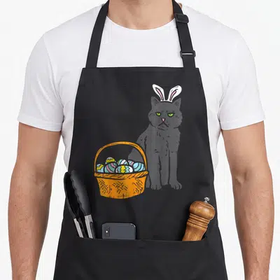 Cat basket apron - Grumpy black cat with white bunny ears sitting behind an orange basket filled with colorful easter — shown on a black cotton canvas apron against a white background