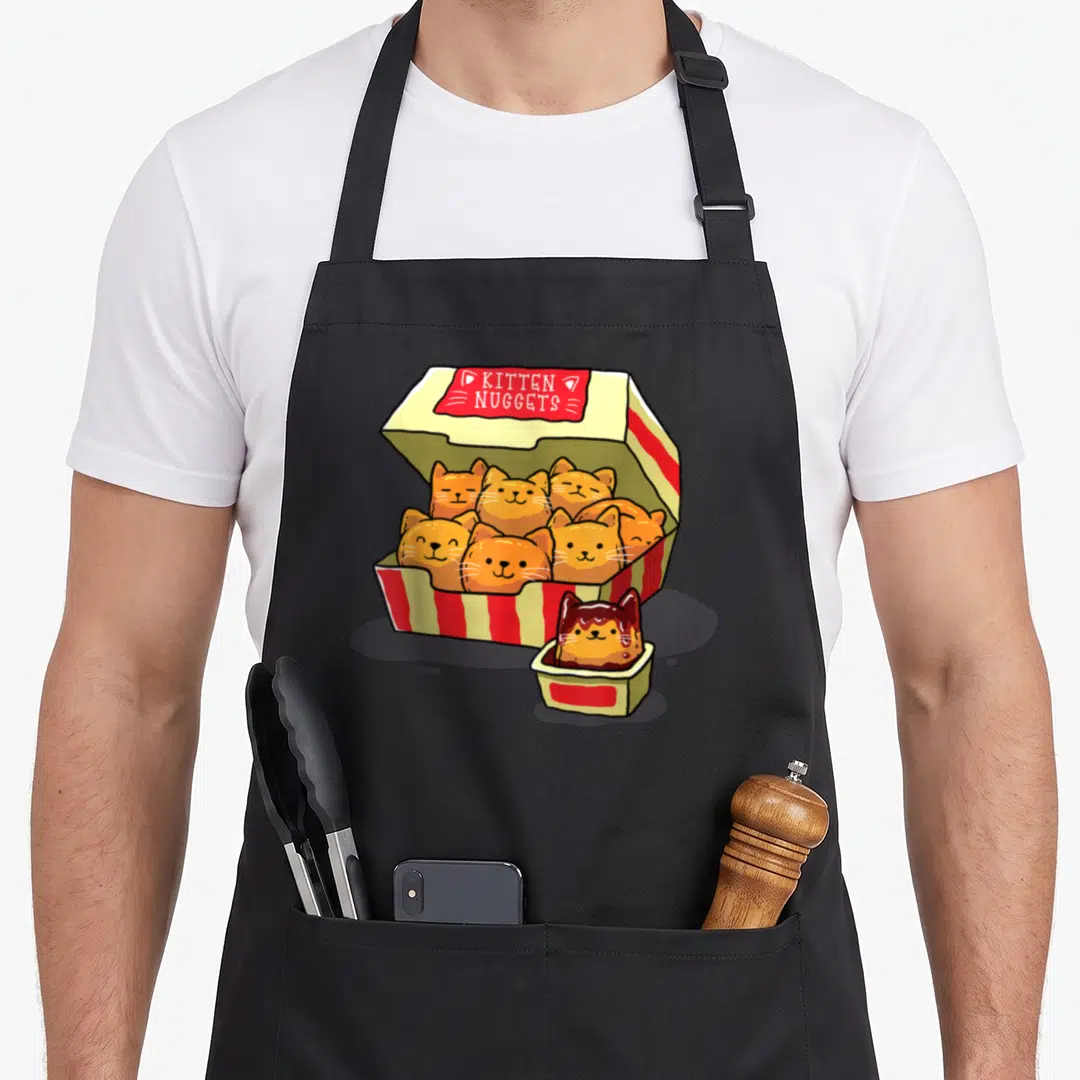 Ingenius funny cat aprons - Cartoon box of orange kitten-shaped nuggets with red sauce cup — shown on a black cotton canvas apron against a white background