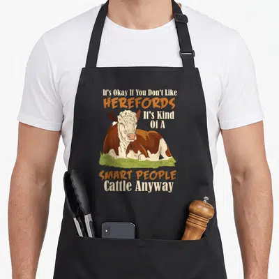 Hereford smart apron - Cartoon cow lying on green grass with white and brown patches and orange text — shown on a black cotton canvas apron against a white background