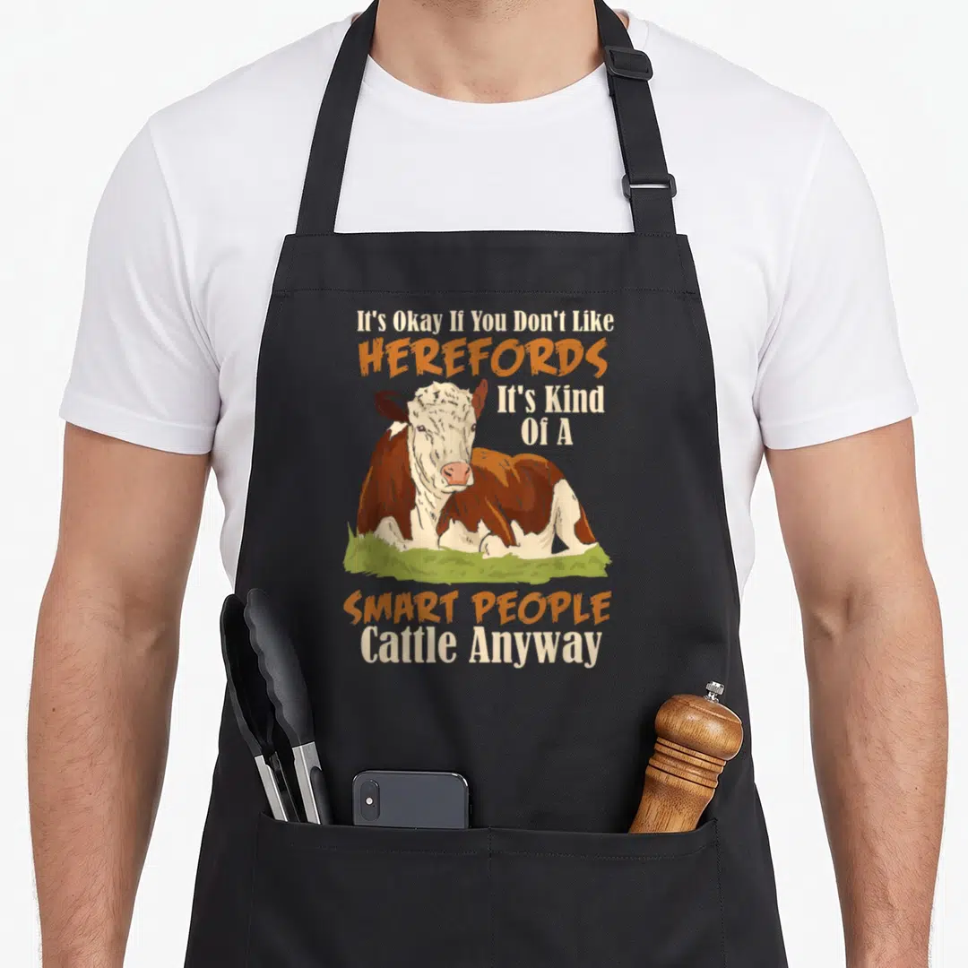 Hereford smart apron - Cartoon cow lying on green grass with white and brown patches and orange text — shown on a black cotton canvas apron against a white background
