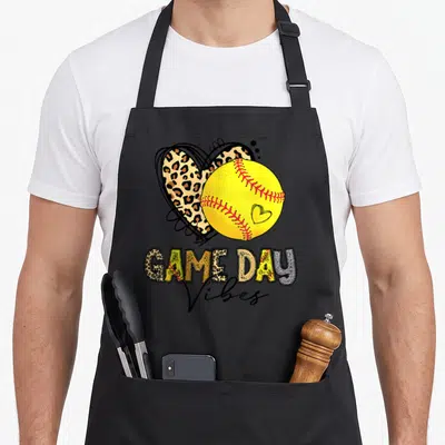 Bleached leopard print cooking apron - Yellow softball overlaps leopard heart with GAME DAY Vibes text in cartoon style — shown on a black cotton canvas apron against a white background