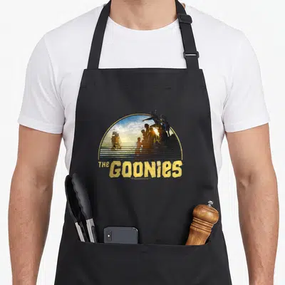 Ship pirate apron - Retro semi-circle with The Goonies text, child silhouettes, and ship against sunset in yellow — shown on a black cotton canvas apron against a white background