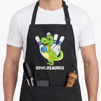 Bowling dinosaur apron - Green cartoon dinosaur holding blue ball in front of four white pins with BOWLOSAURUS text — shown on a black cotton canvas apron against a white background