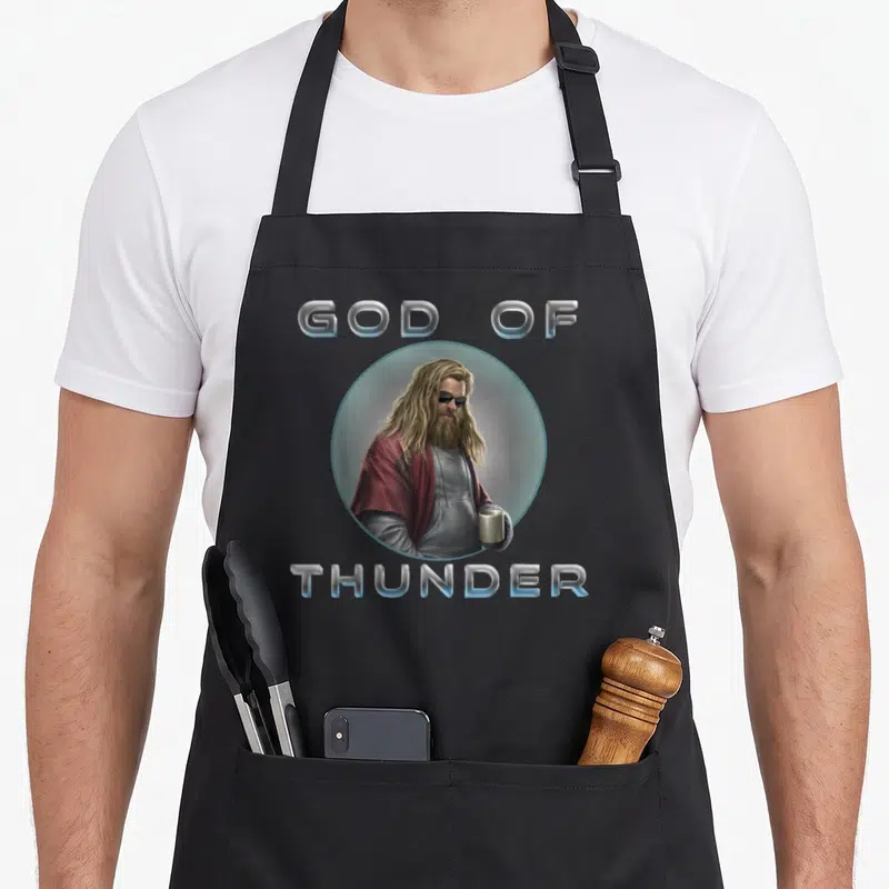 Endgame thunder apron - Man with blonde hair and sunglasses holding a silver mug inside a blue circle with GOD — shown on a black cotton canvas apron against a white background