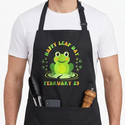 Leapling leap apron - Cartoon frog sitting on lily pad with Happy Leap Day text and footprints — shown on a black cotton canvas apron against a white background