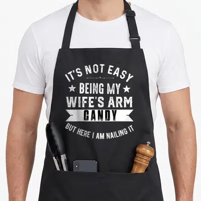 Easy wife arm candy father day apron - Distressed white and black text reading IT'S NOT EASY BEING MY WIFE'S ARM CANDY — shown on a black cotton canvas apron against a white background