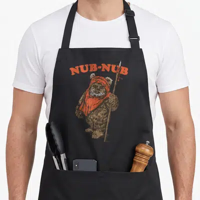 Nub writing lover apron - Brown bear with orange scarf holding spear under arched NUB-NUB text — shown on a black cotton canvas apron against a white background