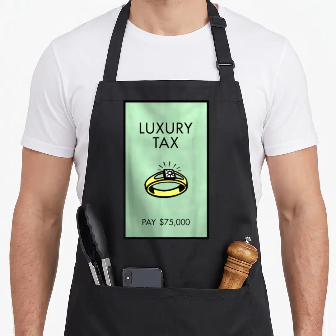 Monopoly luxury tax apron - Light green card with black text reading Luxury Tax Pay 75000 surrounding a yellow ring — shown on a black cotton canvas apron against a white background