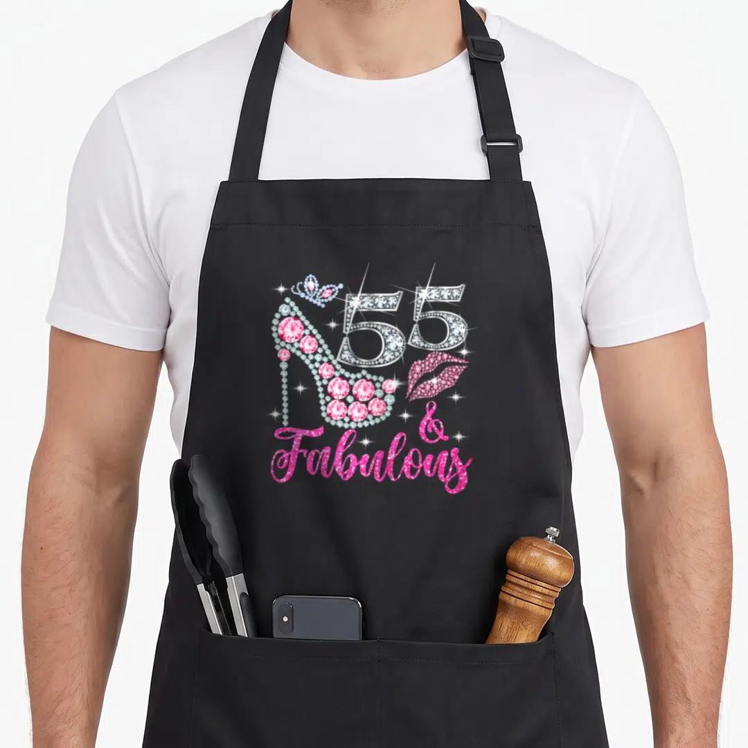 Fabulous queen of baking apron - Diamond encrusted high heel, number 55, pink lips, and text reading 55 & Fabulous — shown on a black cotton canvas apron against a white background