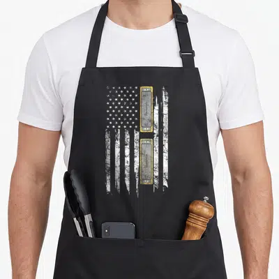 Harmonica patriotic apron - Two stacked harmonicas labeled C and G on a distressed white and grey USA flag background — shown on a black cotton canvas apron against a white background