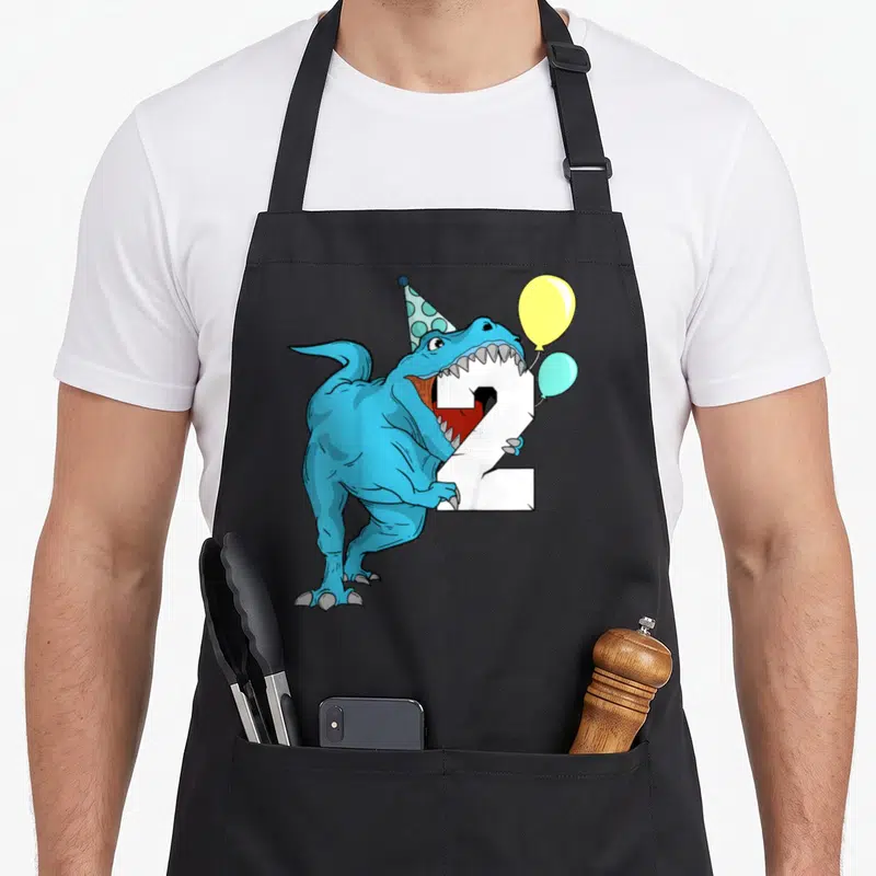 Dinosaur 2nd t-rex apron - Blue T-Rex biting white number 2, wearing polka dot party hat, two balloons yellow and light — shown on a black cotton canvas apron against a white background