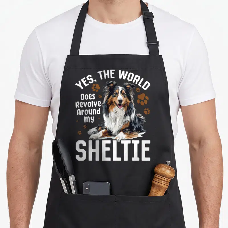 Revolve dog apron - Realistic Shetland Sheepdog illustration with Yes The World Does Revolve Around My Sheltie text — shown on a black cotton canvas apron against a white background