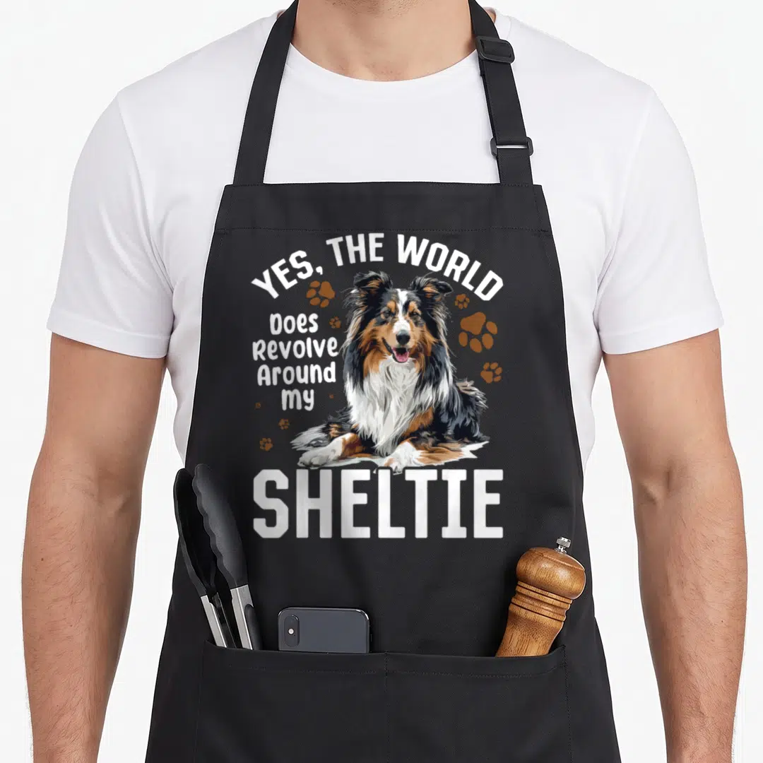 Revolve dog apron - Realistic Shetland Sheepdog illustration with Yes The World Does Revolve Around My Sheltie text — shown on a black cotton canvas apron against a white background