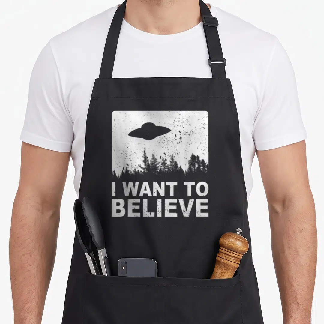 Believe alien ufo apron - Grey UFO silhouette over forest line in white distressed square frame with text I WANT — shown on a black cotton canvas apron against a white background