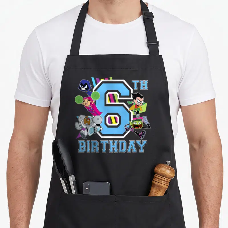 Cute 6th birthday apron - Large blue number 6 with five cartoon characters and birthday text in purple and pink — shown on a black cotton canvas apron against a white background