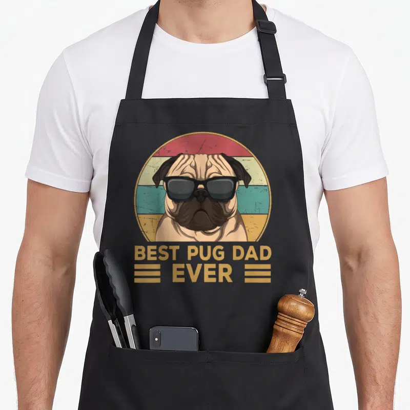 Pug dog dad apron - Retro pug in sunglasses inside gold red and teal striped circle with bold text — shown on a black cotton canvas apron against a white background