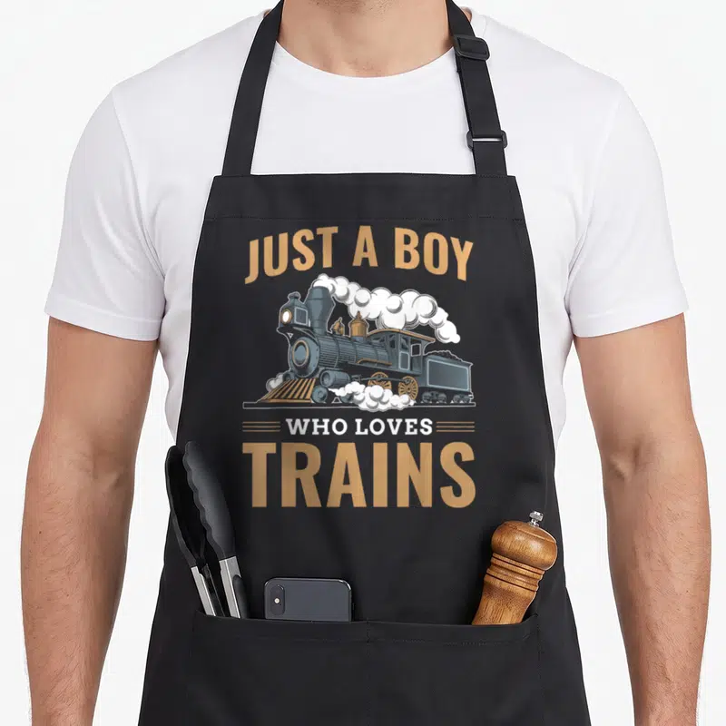 Railroad boy train apron - Retro steam locomotive with arched tan text reading JUST A BOY WHO LOVES TRAINS in blue grey — shown on a black cotton canvas apron against a white background