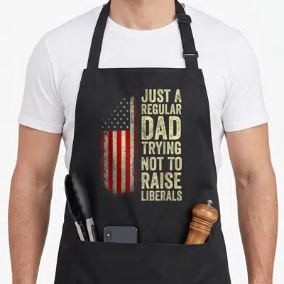 Trying father day apron - Distressed USA flag with stacked beige text reading Just A Regular Dad Trying Not To Raise — shown on a black cotton canvas apron against a white background