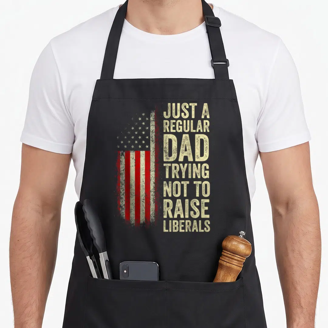 Trying father day apron - Distressed USA flag with stacked beige text reading Just A Regular Dad Trying Not To Raise — shown on a black cotton canvas apron against a white background