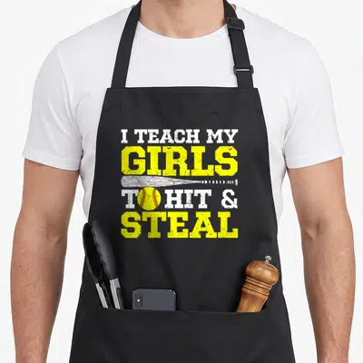 Teach softball mom apron - Stacked text I teach my girls to hit and steal with white bat and yellow ball — shown on a black cotton canvas apron against a white background