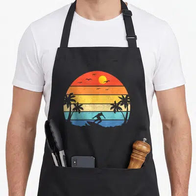 Beach surfing player apron - Retro sunset with striped colors and grey silhouettes of a surfer, palms, and birds — shown on a black cotton canvas apron against a white background