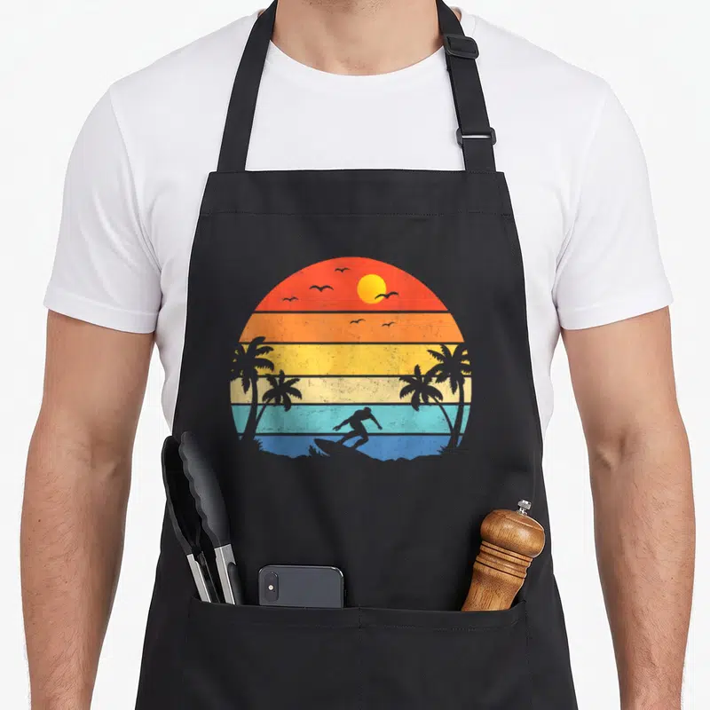 Beach surfing player apron - Retro sunset with striped colors and grey silhouettes of a surfer, palms, and birds — shown on a black cotton canvas apron against a white background