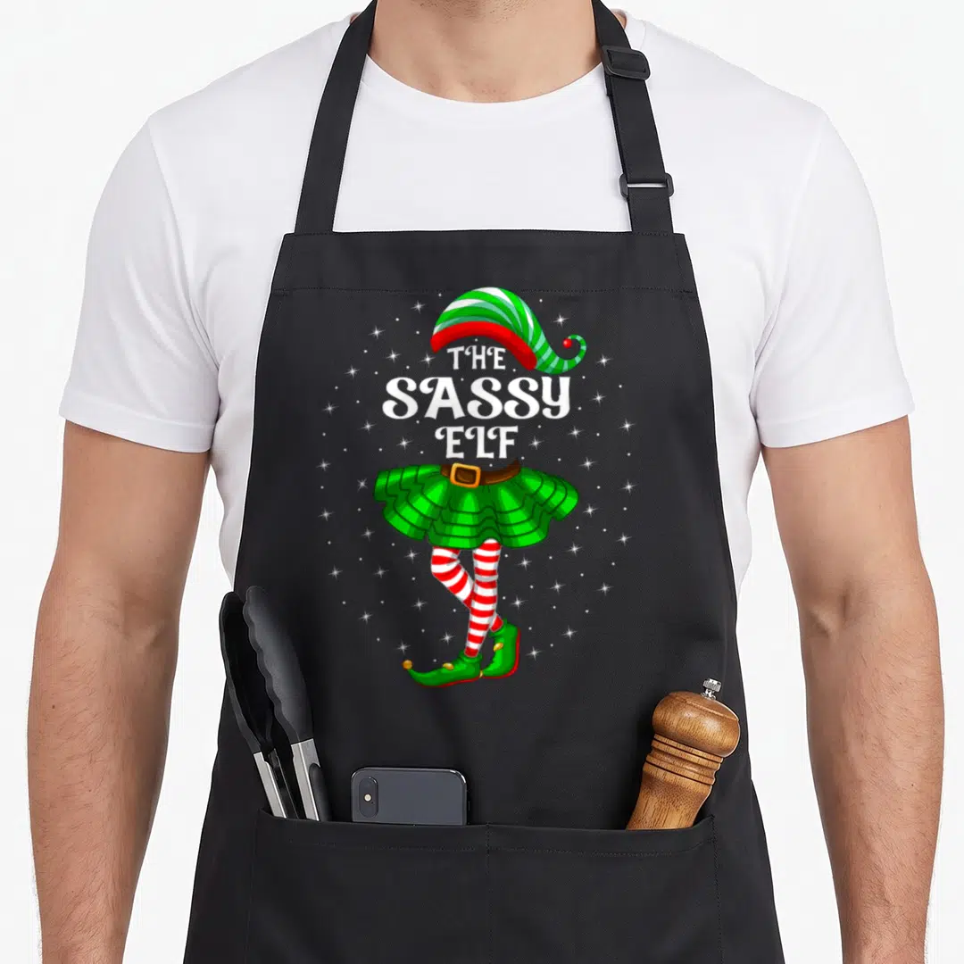 Xmas family christmas elf apron - Cartoon elf with green skirt and striped legs under The Sassy Elf text surrounded — shown on a black cotton canvas apron against a white background