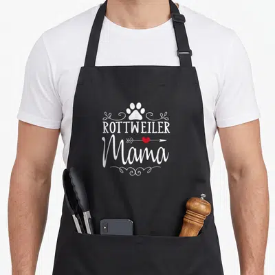 Rottweiler mama apron - White paw print above ROTTWEILER Mama text with red heart arrow and swirls — shown on a black cotton canvas apron against a white background