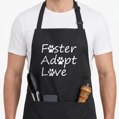 Foster adopt grill rescue apron - White text Foster Adopt Love with paw prints replacing the o letters — shown on a black cotton canvas apron against a white background