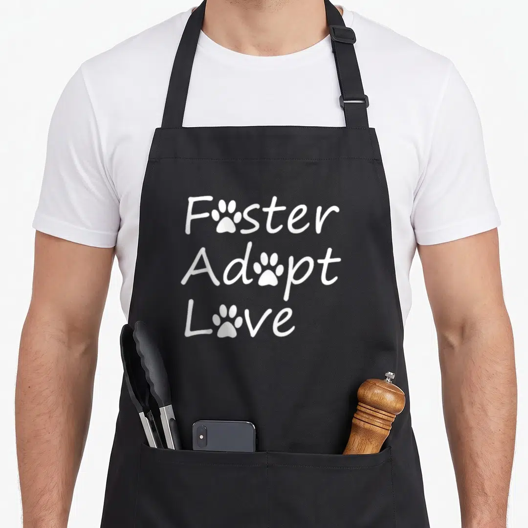 Foster adopt grill rescue apron - White text Foster Adopt Love with paw prints replacing the o letters — shown on a black cotton canvas apron against a white background