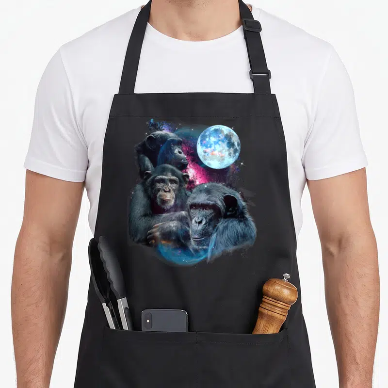 Chimp animal apron - Three realistic chimpanzees clustered near a blue moon and pink cosmic nebula — shown on a black cotton canvas apron against a white background
