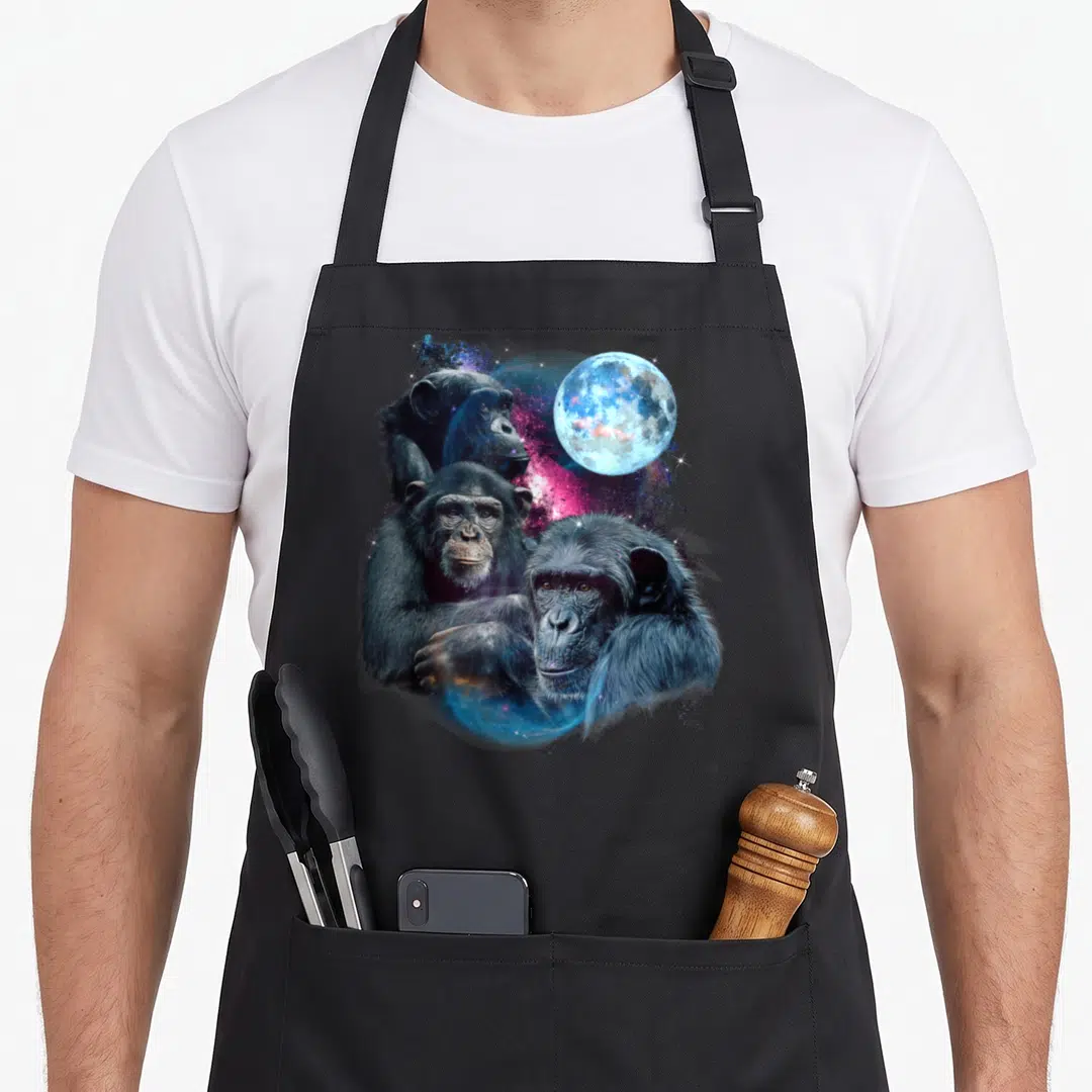 Chimp animal apron - Three realistic chimpanzees clustered near a blue moon and pink cosmic nebula — shown on a black cotton canvas apron against a white background