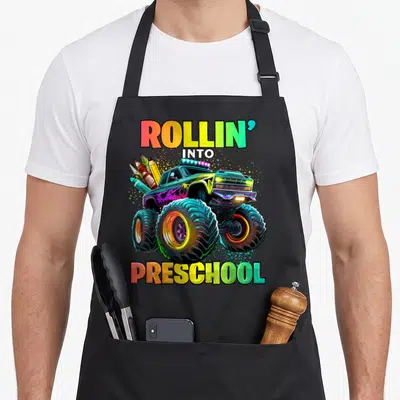 Preschool monster truck apron - Cartoon monster truck carrying pencils and crayons with ROLLIN INTO PRESCHOOL text — shown on a black cotton canvas apron against a white background