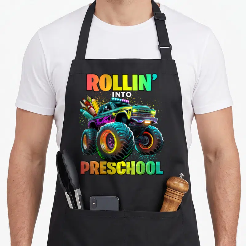 Preschool monster truck apron - Cartoon monster truck carrying pencils and crayons with ROLLIN INTO PRESCHOOL text — shown on a black cotton canvas apron against a white background
