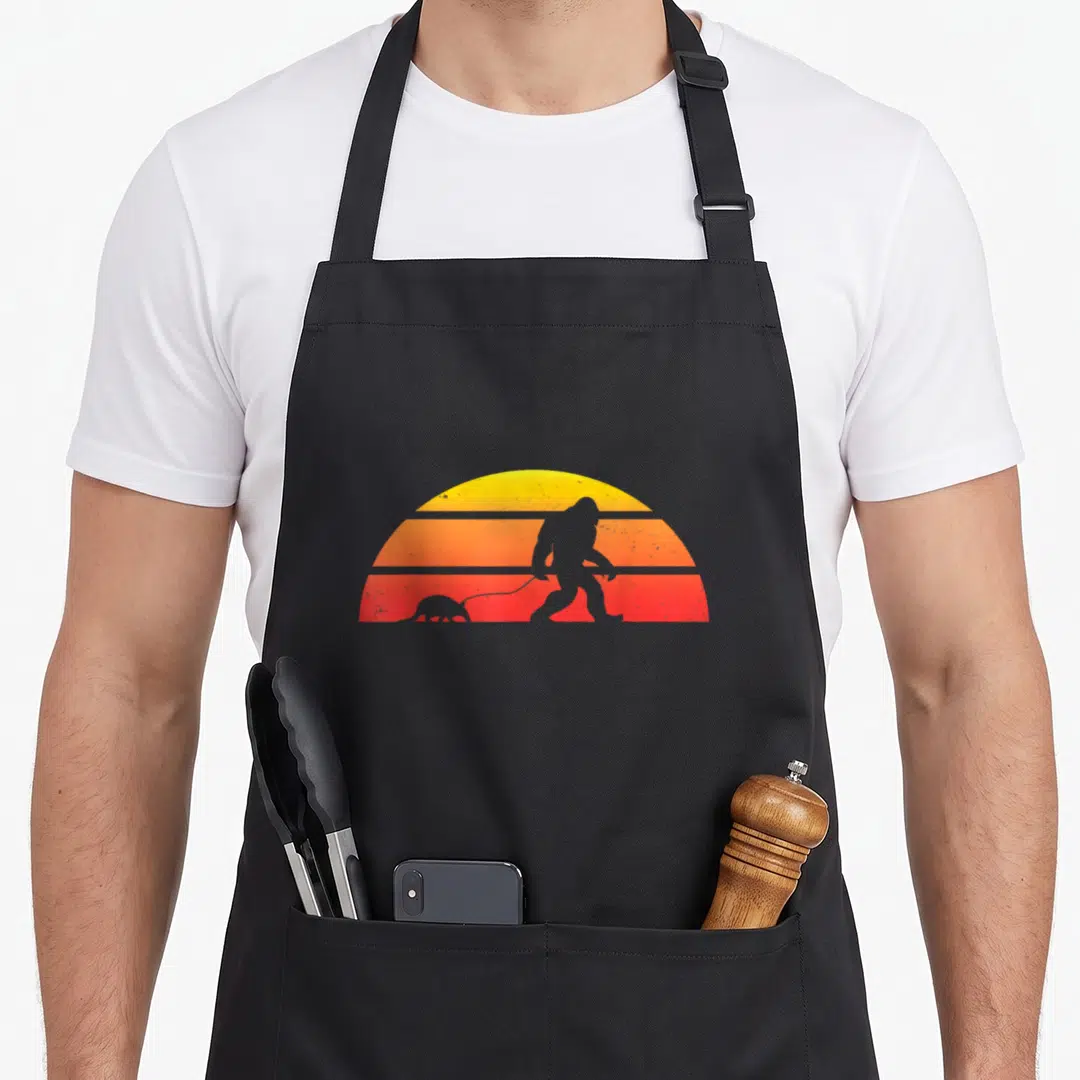 Armadillo bigfoot apron - Grey Bigfoot silhouette walking small animal on leash before striped yellow orange red sun — shown on a black cotton canvas apron against a white background