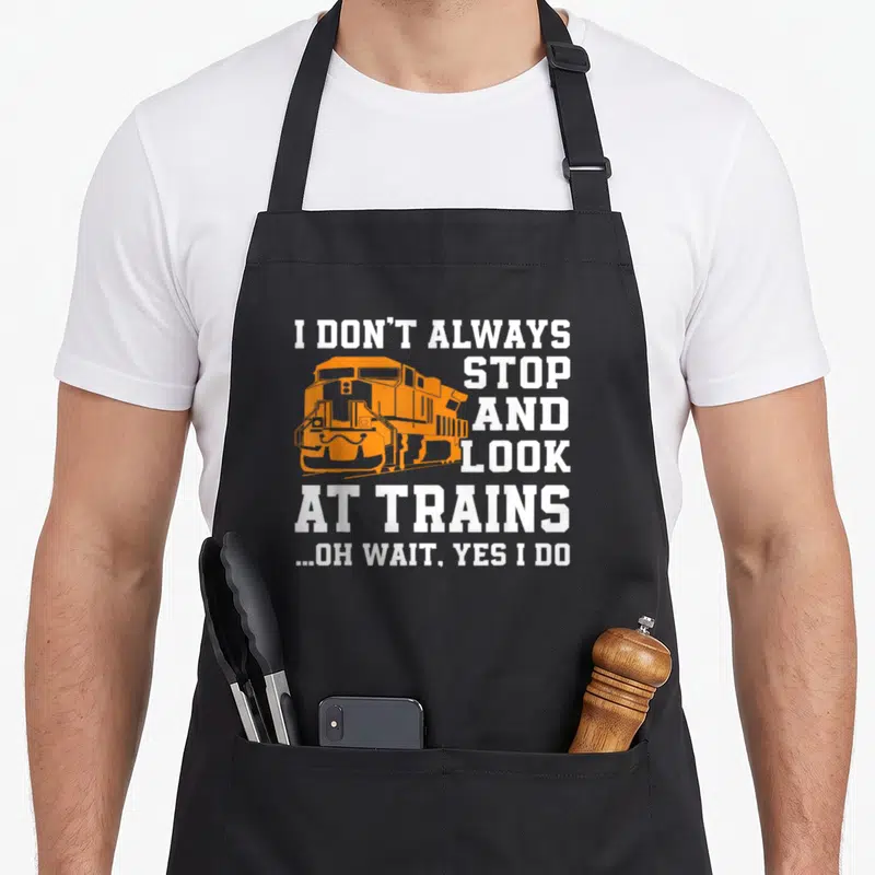 Train locomotive railroad apron - Orange train graphic with white text reading I don't always stop and look at trains — shown on a black cotton canvas apron against a white background