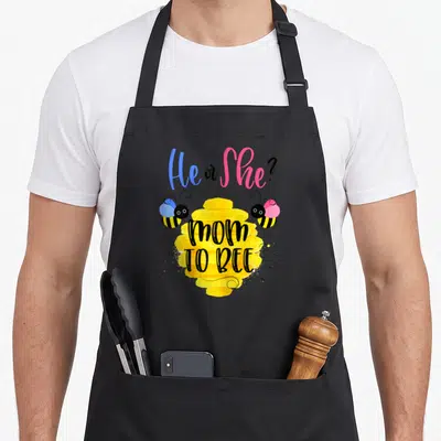 Gender reveal bee apron - Yellow honeycomb with mom TO BEE text flanked by blue and pink bow bees — shown on a black cotton canvas apron against a white background