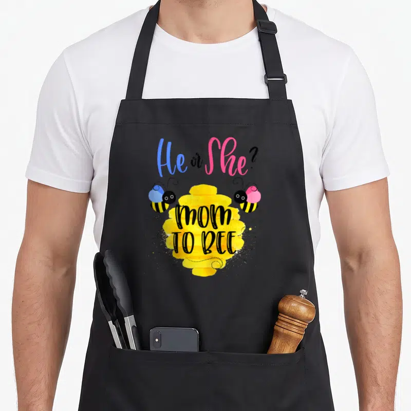 Gender reveal bee apron - Yellow honeycomb with mom TO BEE text flanked by blue and pink bow bees — shown on a black cotton canvas apron against a white background