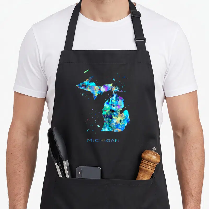 Michigan travel girl apron - Watercolor map of Michigan state in blue, cyan, yellow, purple, and green splashes — shown on a black cotton canvas apron against a white background