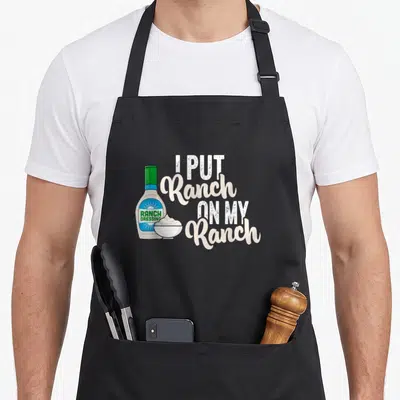 Vintage ranch dressing apron - Cartoon ranch bottle and bowl next to stacked text I Put Ranch On My Ranch in white — shown on a black cotton canvas apron against a white background