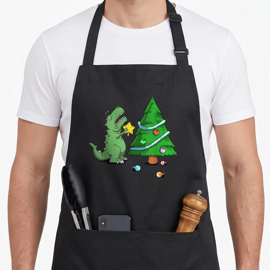 Struggle christmas tree apron - Green dinosaur holding yellow star next to Christmas tree with scattered ornaments — shown on a black cotton canvas apron against a white background