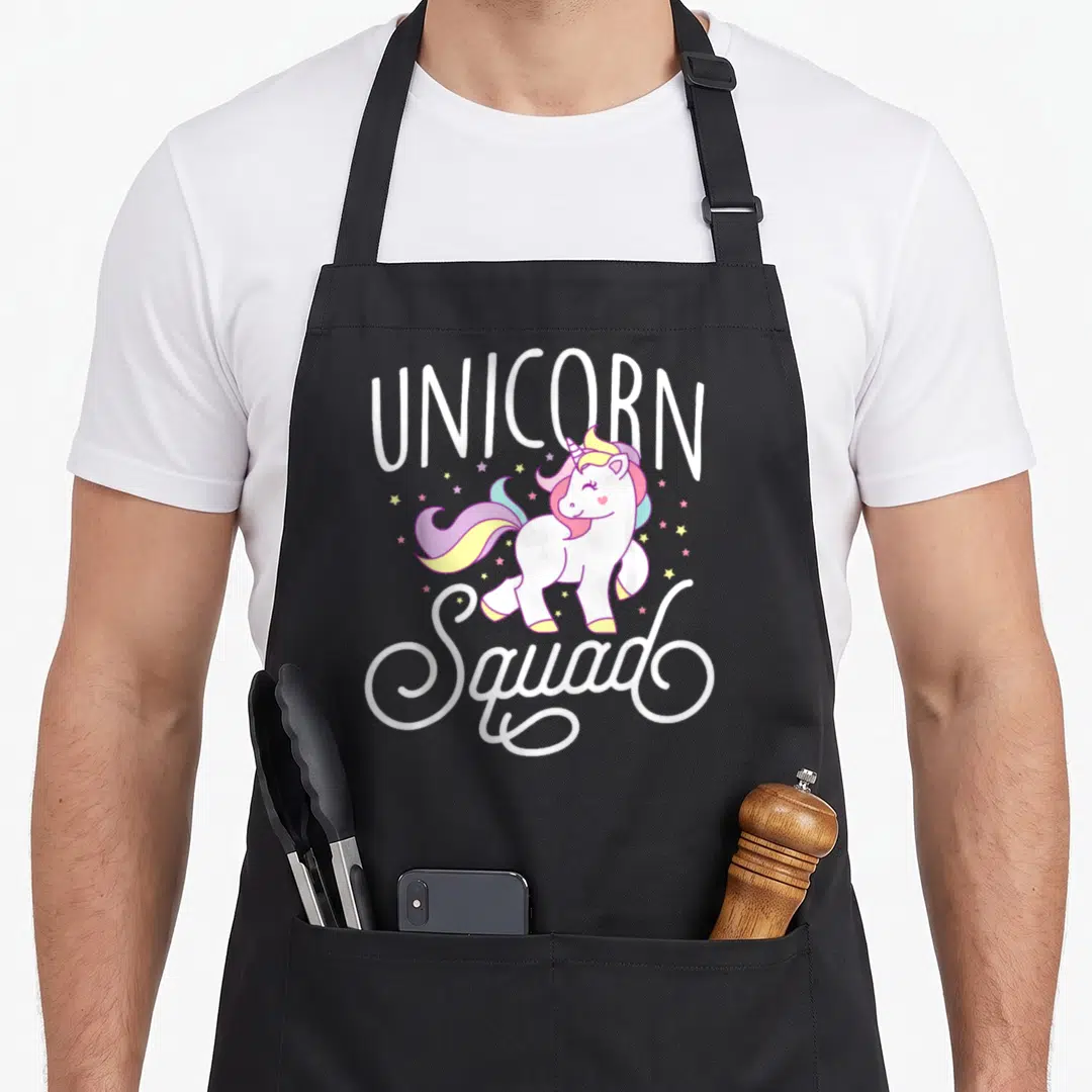 Cute squad unicorn apron - Arched UNICORN and Squad text framing a cartoon white unicorn with pink mane and scattered — shown on a black cotton canvas apron against a white background