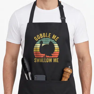Swallow turkey apron - GOBBLE ME SWALLOW ME text over striped retro sun with grey turkey silhouette — shown on a black cotton canvas apron against a white background